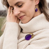 A woman wearing a cream-colored turtleneck sweater stands outdoors, smiling gently with eyes closed. She has wavy hair and wears matching purple jewelry—a handcrafted brooch and earrings. The grassy background is slightly blurred.