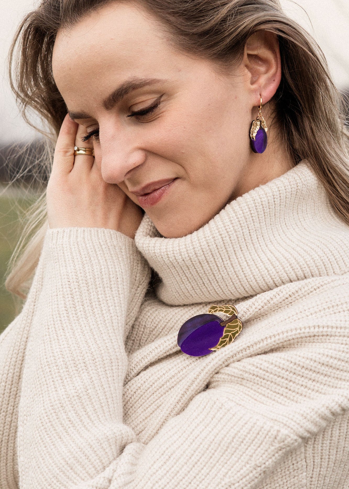 A woman wearing a cream-colored turtleneck sweater stands outdoors, smiling gently with eyes closed. She has wavy hair and wears matching purple jewelry—a handcrafted brooch and earrings. The grassy background is slightly blurred.