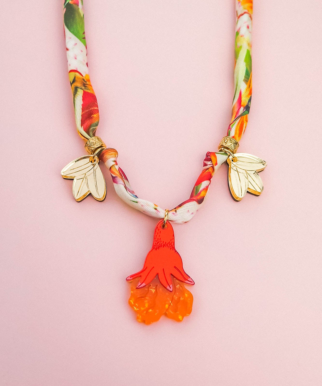 A colorful handmade necklace with a floral-patterned fabric band, two gold flower charms, and a central pendant shaped like an orange and red flower—perfect as a unique pomegranate flower necklace—displayed on a light pink background.