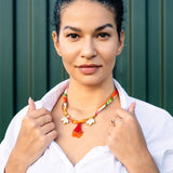 A woman with dark hair in a bun wears a white shirt and a handmade pomegranate flower necklace featuring floral charms on a silk-print cord, standing confidently before a green striped background while holding her collar.