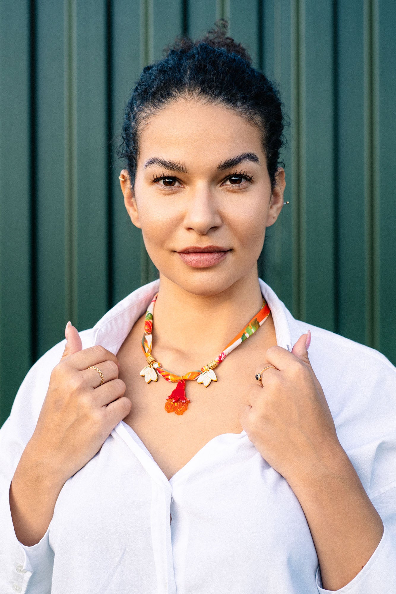A woman with dark hair in a bun wears a white shirt and a handmade pomegranate flower necklace featuring floral charms on a silk-print cord, standing confidently before a green striped background while holding her collar.