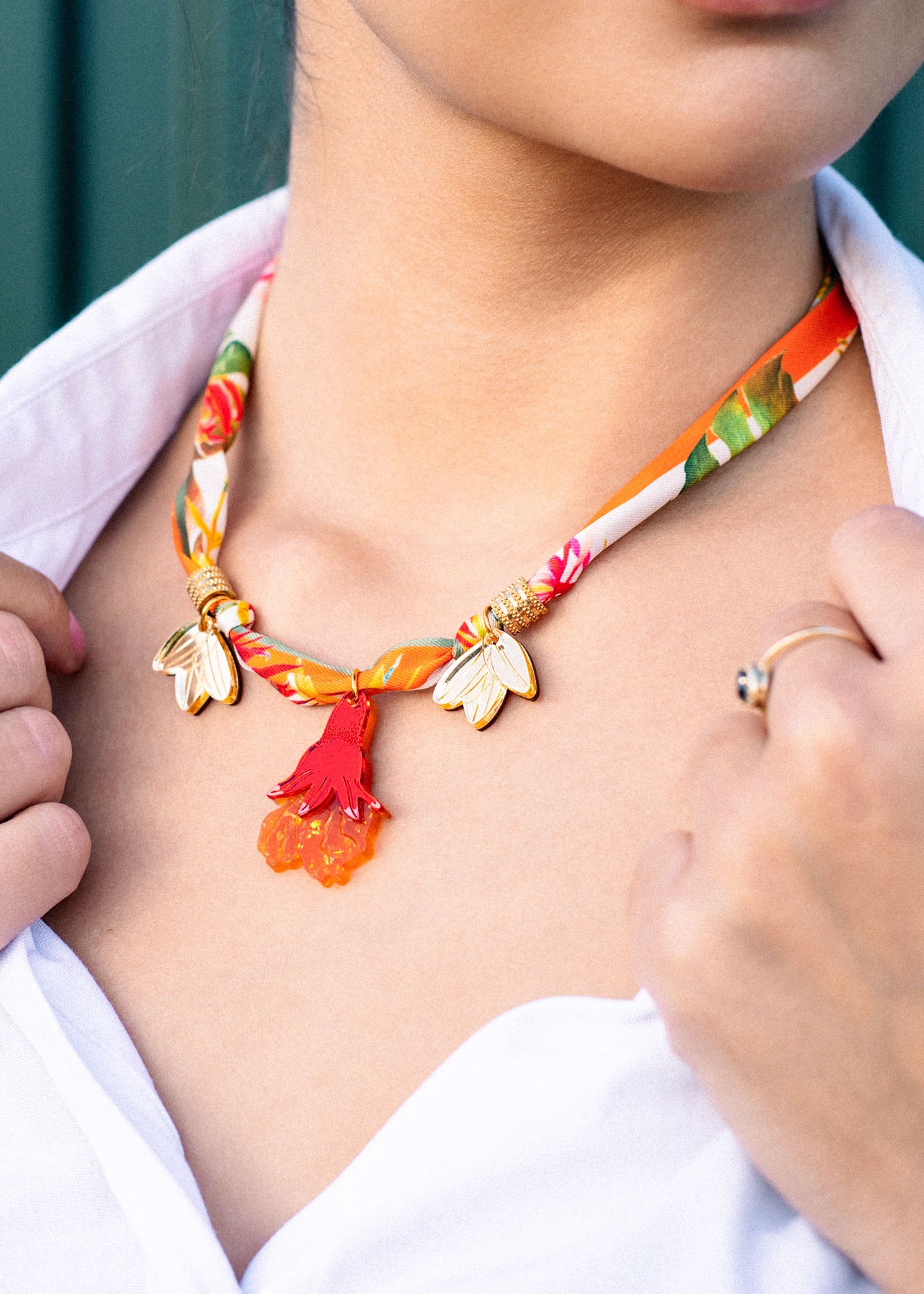 A woman wearing a handmade pomegranate flower necklace with gold accents, red tassels, and orange flower-shaped charms, paired with a white collared shirt. Only her lower face, neck, and hand are visible.