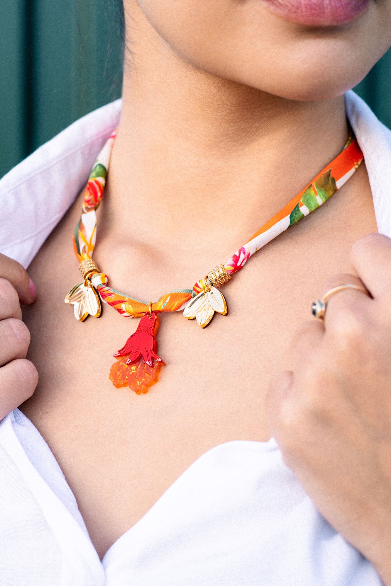 A woman wearing a handmade pomegranate flower necklace with gold accents, red tassels, and orange flower-shaped charms, paired with a white collared shirt. Only her lower face, neck, and hand are visible.