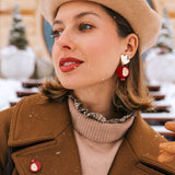 A woman in a tan beret and brown coat stands outside on a snowy day, wearing bold, handcrafted earrings with a bird design, a matching pin, tan gloves, and red lipstick. Snowflakes sprinkle her hair and coat.