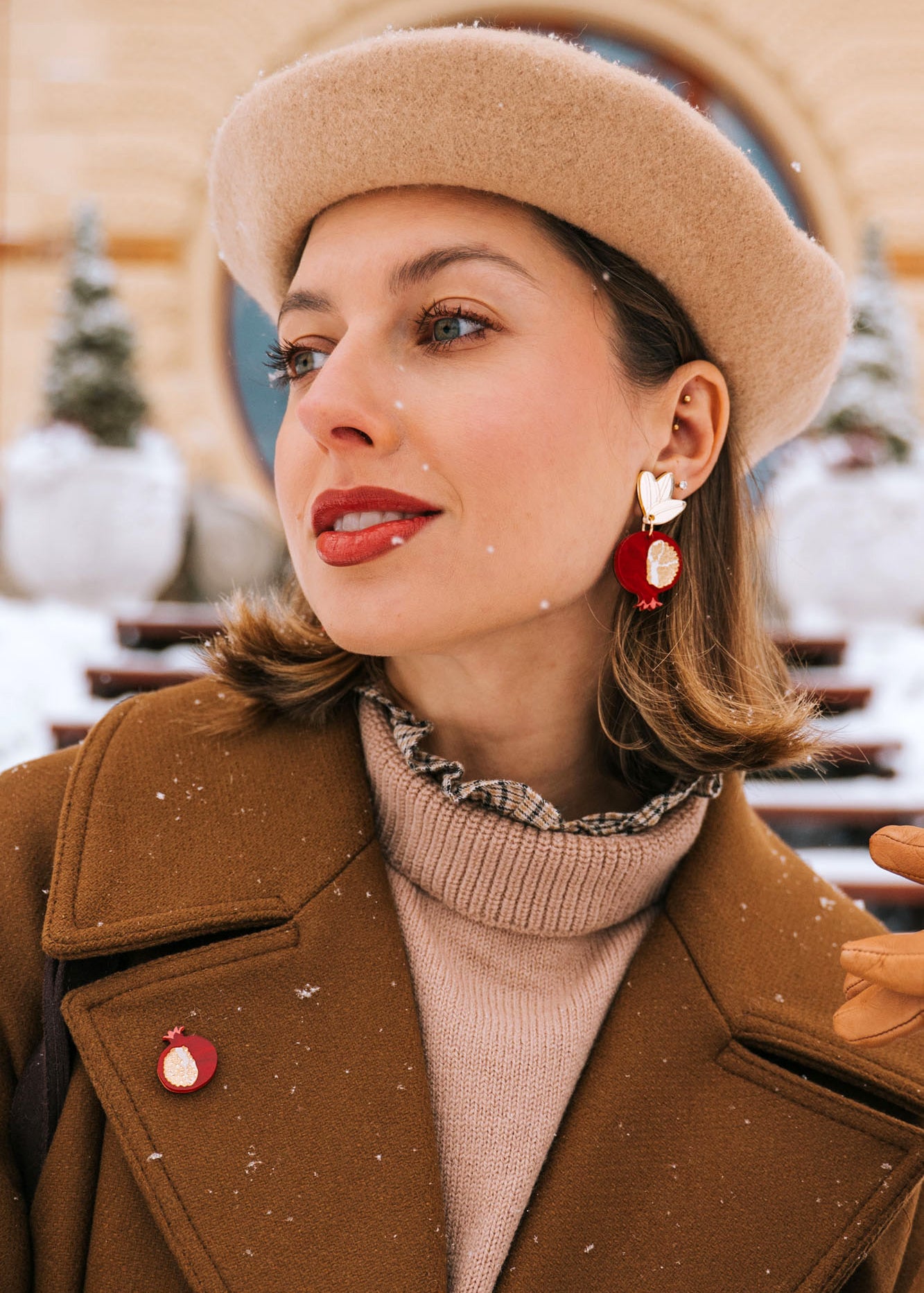 A woman in a tan beret and brown coat stands outside on a snowy day, wearing bold, handcrafted earrings with a bird design, a matching pin, tan gloves, and red lipstick. Snowflakes sprinkle her hair and coat.