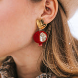 A close-up of a woman's ear wearing a gold stud and a large, handcrafted pomegranate-shaped earring with black leaves. She has a beige beret and a brown coat with a plaid collar. Snowflakes rest on her hair and coat, highlighting bold designs.