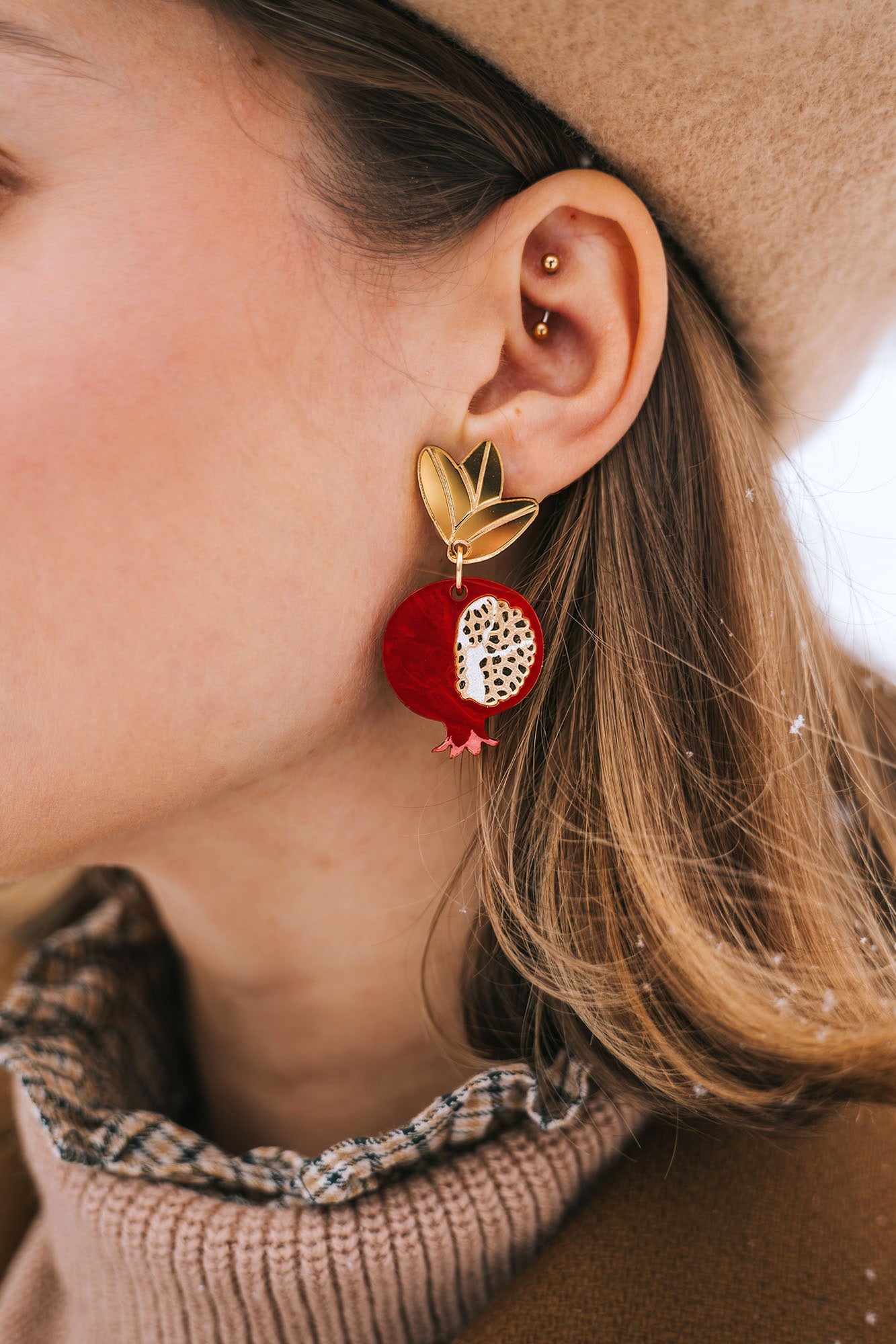 A close-up of a woman's ear wearing a gold stud and a large, handcrafted pomegranate-shaped earring with black leaves. She has a beige beret and a brown coat with a plaid collar. Snowflakes rest on her hair and coat, highlighting bold designs.