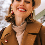 A woman wearing a beige beret, brown coat, and handcrafted earrings with red accents smiles outdoors on a snowy day. Snow is visible on her coat and in the background.