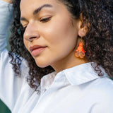 A woman with curly hair wearing a white collared shirt and handmade floral earrings looks down with a calm expression, her hand resting on her head.