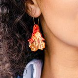 Close-up of a person with curly hair wearing a large, colorful handmade earring shaped like an orange and cream jellyfish. The person is dressed in a light blue collared shirt.