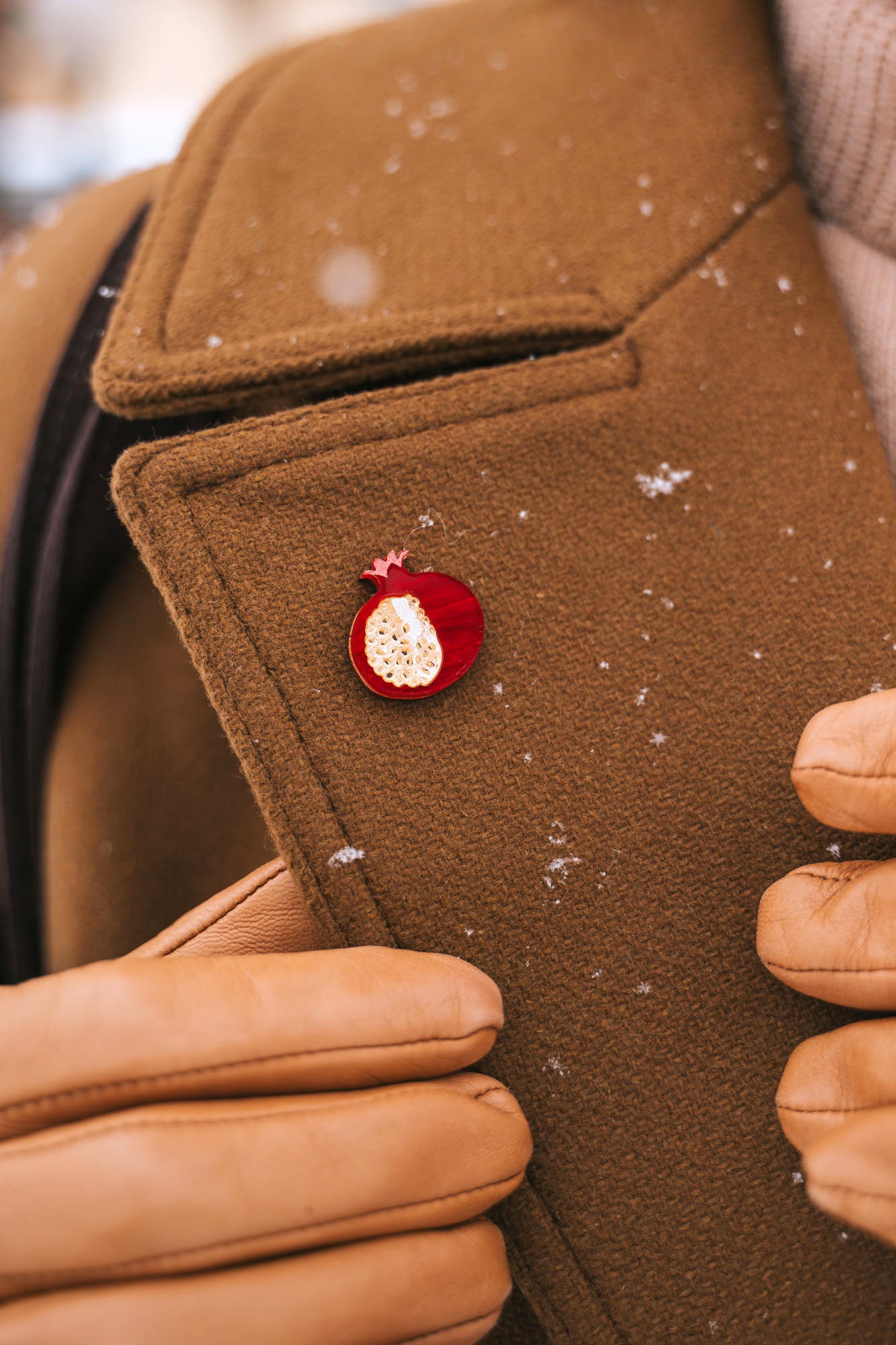 A person wearing a brown coat and tan gloves adjusts the lapel, which is decorated with a handcrafted Pomegranate Pin—a unique fashion accessory. Snowflakes are visible on the coat.
