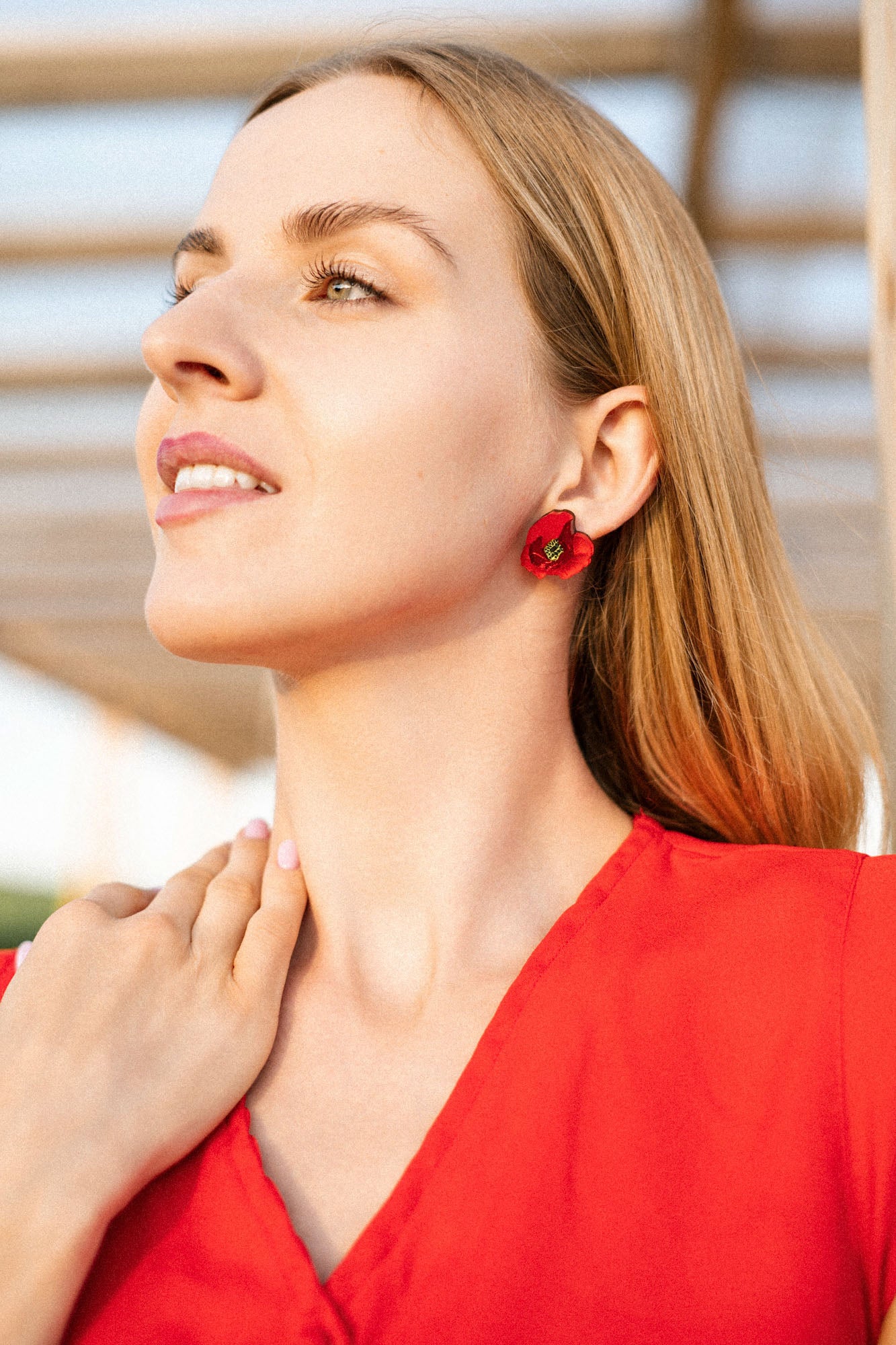 A woman with long blonde hair wears a red top and handcrafted, poppy design acrylic earrings. She looks upward and smiles softly, touching her neck with one hand. The background is softly blurred with sunlight.