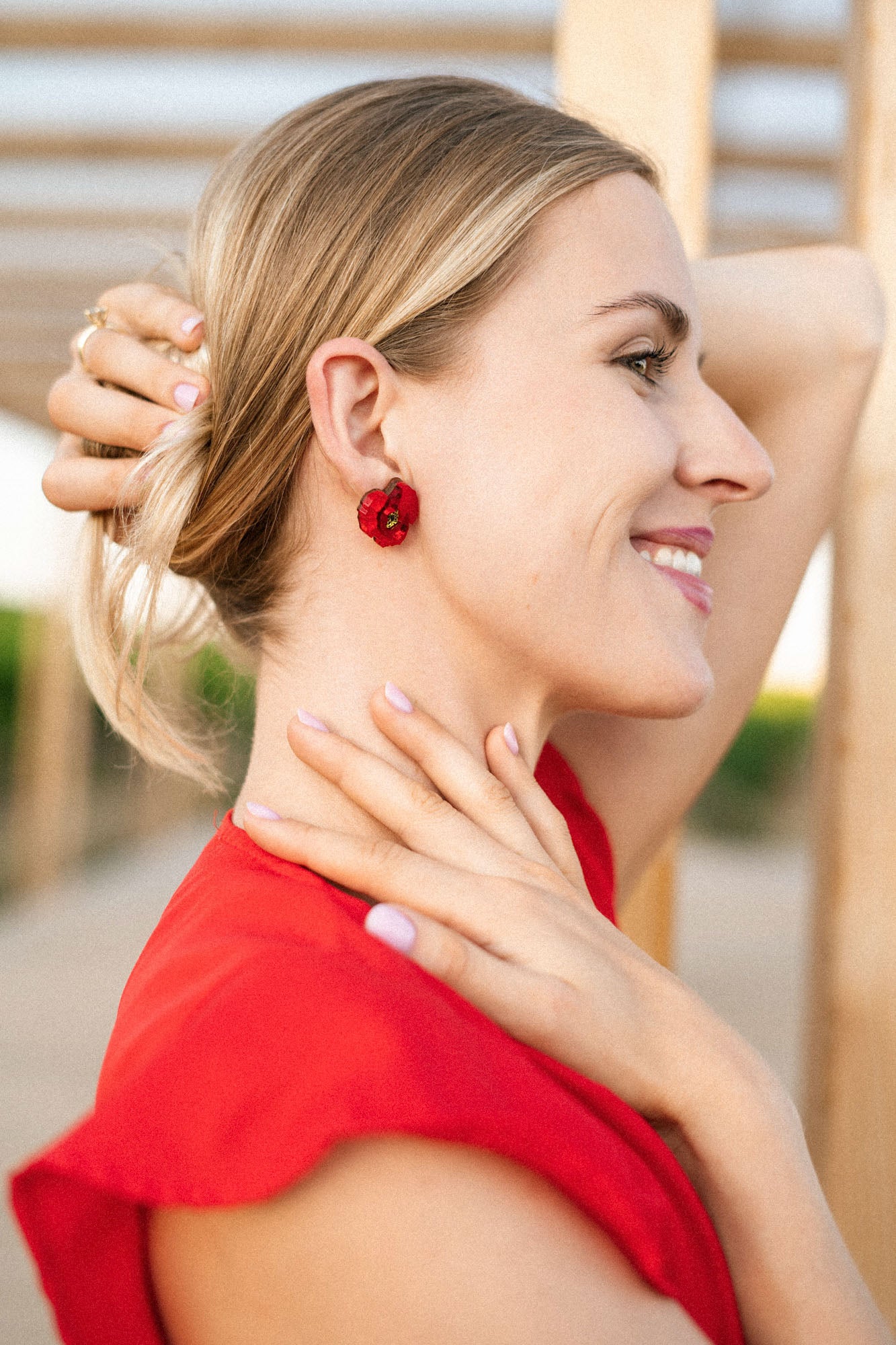 A smiling woman in a red top with handcrafted red flower earrings holds her hair back with one hand and touches her neck with the other. She is outdoors, with wooden beams and greenery in the background.