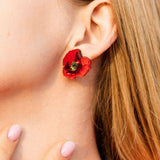 Close-up of a woman’s ear wearing a handcrafted red poppy design acrylic earring. Her blonde hair is visible, and she has light pink nail polish on her fingernails, gently touching her neck.