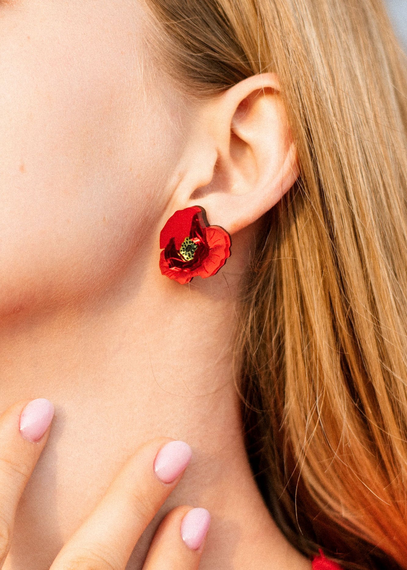 Close-up of a woman’s ear wearing a handcrafted red poppy design acrylic earring. Her blonde hair is visible, and she has light pink nail polish on her fingernails, gently touching her neck.