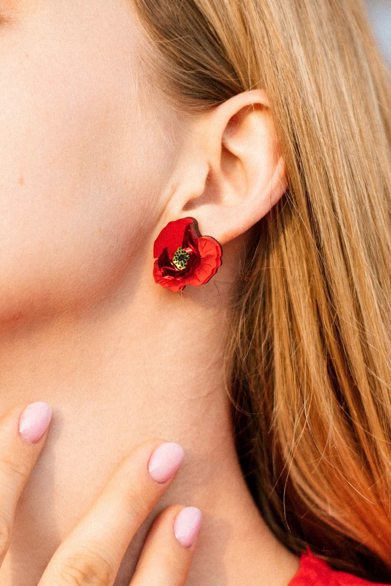 Close-up of a woman’s ear wearing a handcrafted red poppy design acrylic earring. Her blonde hair is visible, and she has light pink nail polish on her fingernails, gently touching her neck.