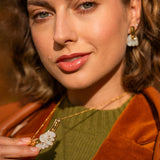 A woman with wavy brown hair wears an elegant green top, brown jacket, and handcrafted gold jewelry with matching white flower-shaped earrings and a Tulip Necklace, holding the necklace and looking confidently at the camera.