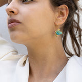 A woman with dark hair pulled back holds her hands behind her head, wearing a white blouse and handcrafted Little Turtle Hoop Earrings with green gemstones, looking thoughtfully into the distance against a plain light background.