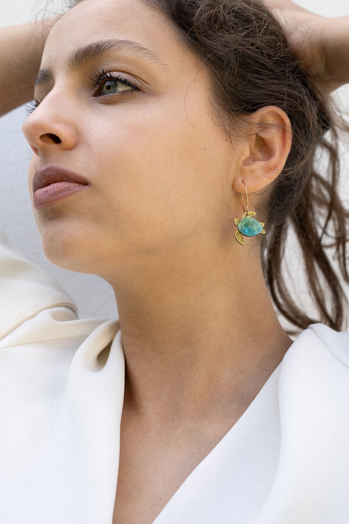A woman with dark hair pulled back holds her hands behind her head, wearing a white blouse and handcrafted Little Turtle Hoop Earrings with green gemstones, looking thoughtfully into the distance against a plain light background.