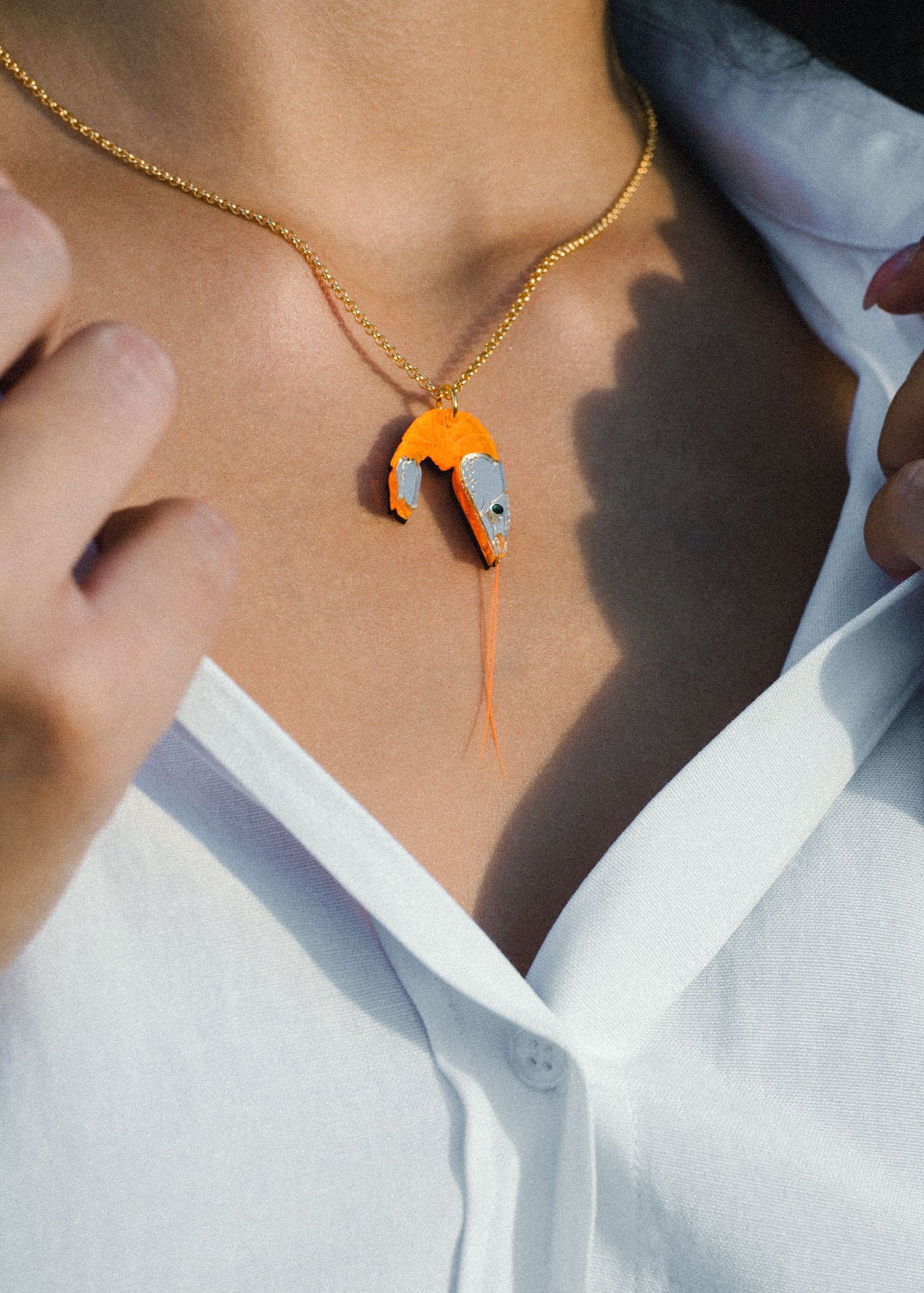 A person wearing a white shirt and a gold playful necklace with a pendant shaped like a shrimp, featuring orange and metallic colors.