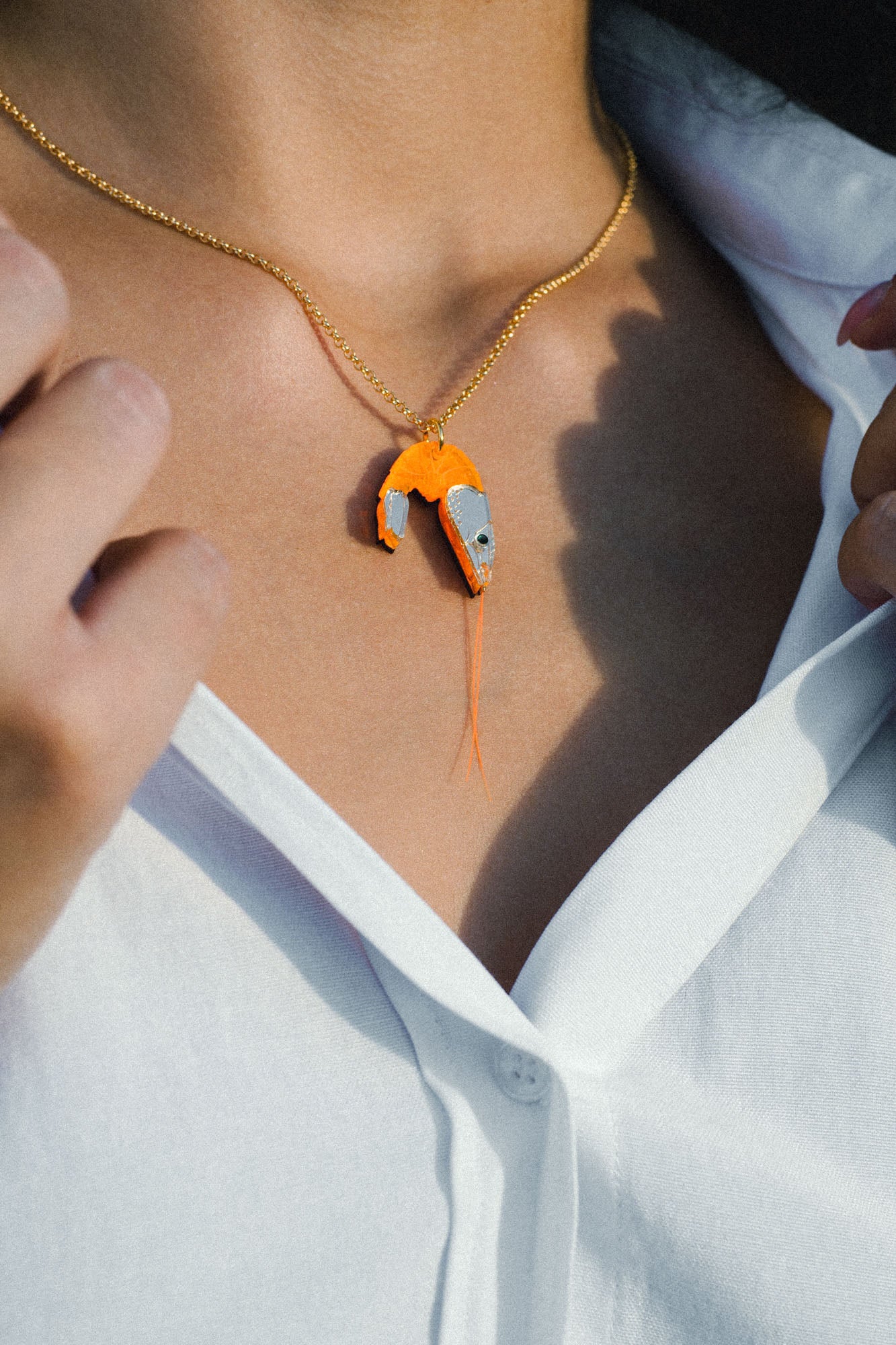 A person wearing a white shirt and a gold playful necklace with a pendant shaped like a shrimp, featuring orange and metallic colors.
