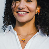 A smiling person with curly hair wears a white shirt and matching orange-and-gold imitation jewelry: a playful necklace and earrings shaped like abstract birds.