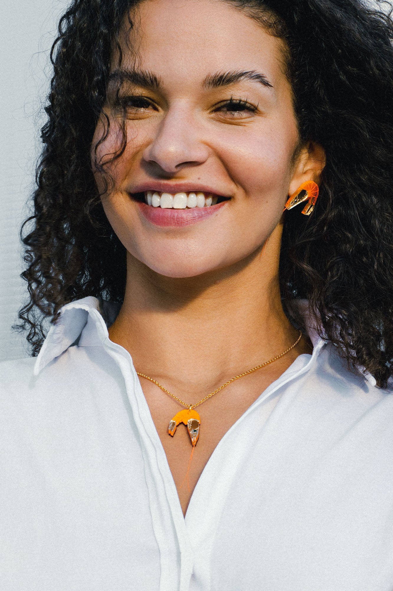 A smiling person with curly hair wears a white shirt and matching orange-and-gold imitation jewelry: a playful necklace and earrings shaped like abstract birds.