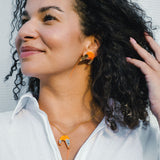 Smiling woman with curly hair wears a white shirt, a playful necklace featuring a shrimp pendant, and matching imitation jewelry earrings. She touches her hair and looks to the side against a light background.