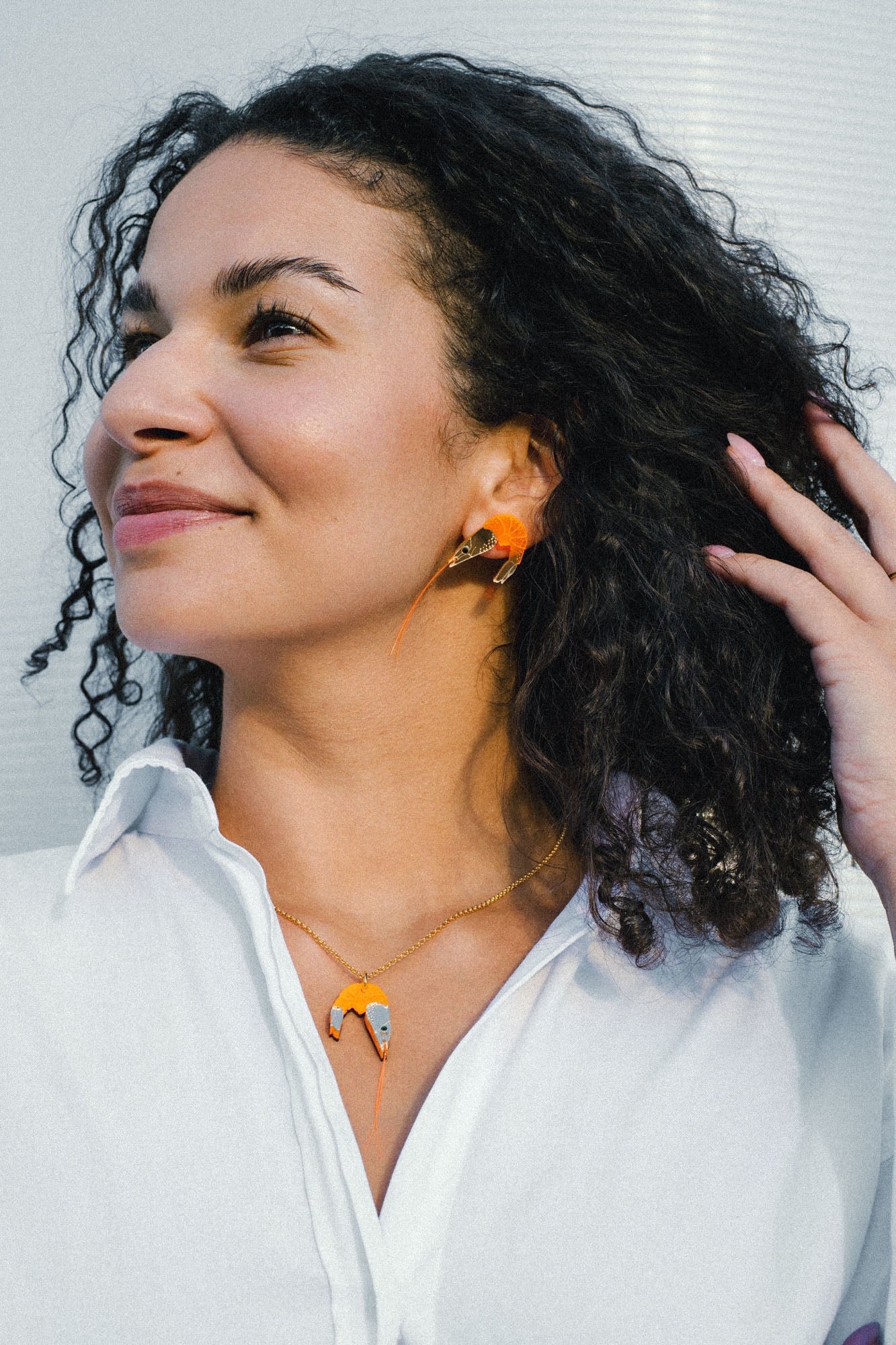 Smiling woman with curly hair wears a white shirt, a playful necklace featuring a shrimp pendant, and matching imitation jewelry earrings. She touches her hair and looks to the side against a light background.