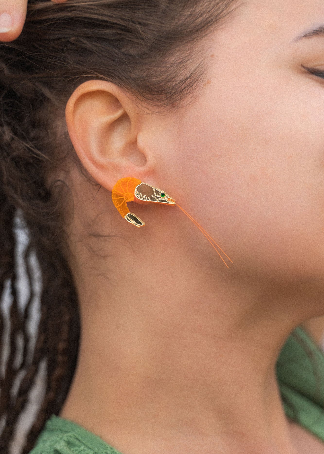 A person with brown skin and dreadlocks wears a detailed, neon orange Shrimp Stud earring adorned with emerald rhinestones. Their hand rests on their head, and they are dressed in a green top.
