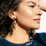 A woman with curly hair wearing a blue top and featherlight gold dangle earrings is shown in close-up, looking to the side in bright, natural light.