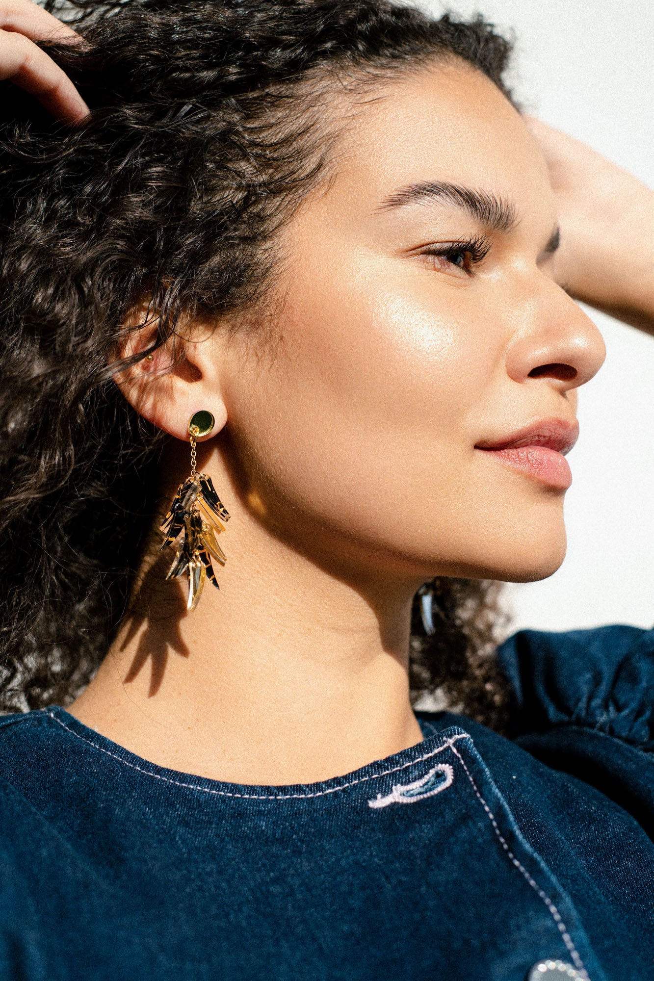 A woman with curly hair wearing a blue top and featherlight gold dangle earrings is shown in close-up, looking to the side in bright, natural light.