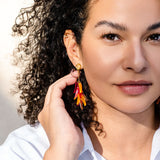 A woman with curly hair in a white shirt holds her hand to her ear, showing off featherlight orange and red dangle earrings. She has a calm expression and natural makeup against a bright, neutral background.
