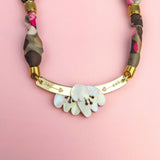 A handmade necklace with a gold curved bar, white shell-like pieces, and a multicolored fabric chain reminiscent of a silk-print scarf in green, pink, brown, and gold accents, displayed on a light pink background.
