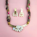 A handmade necklace with a fabric-wrapped chain and matching earrings, both featuring white, abstract, acrylic tulip-like pendants, displayed on a pink background.