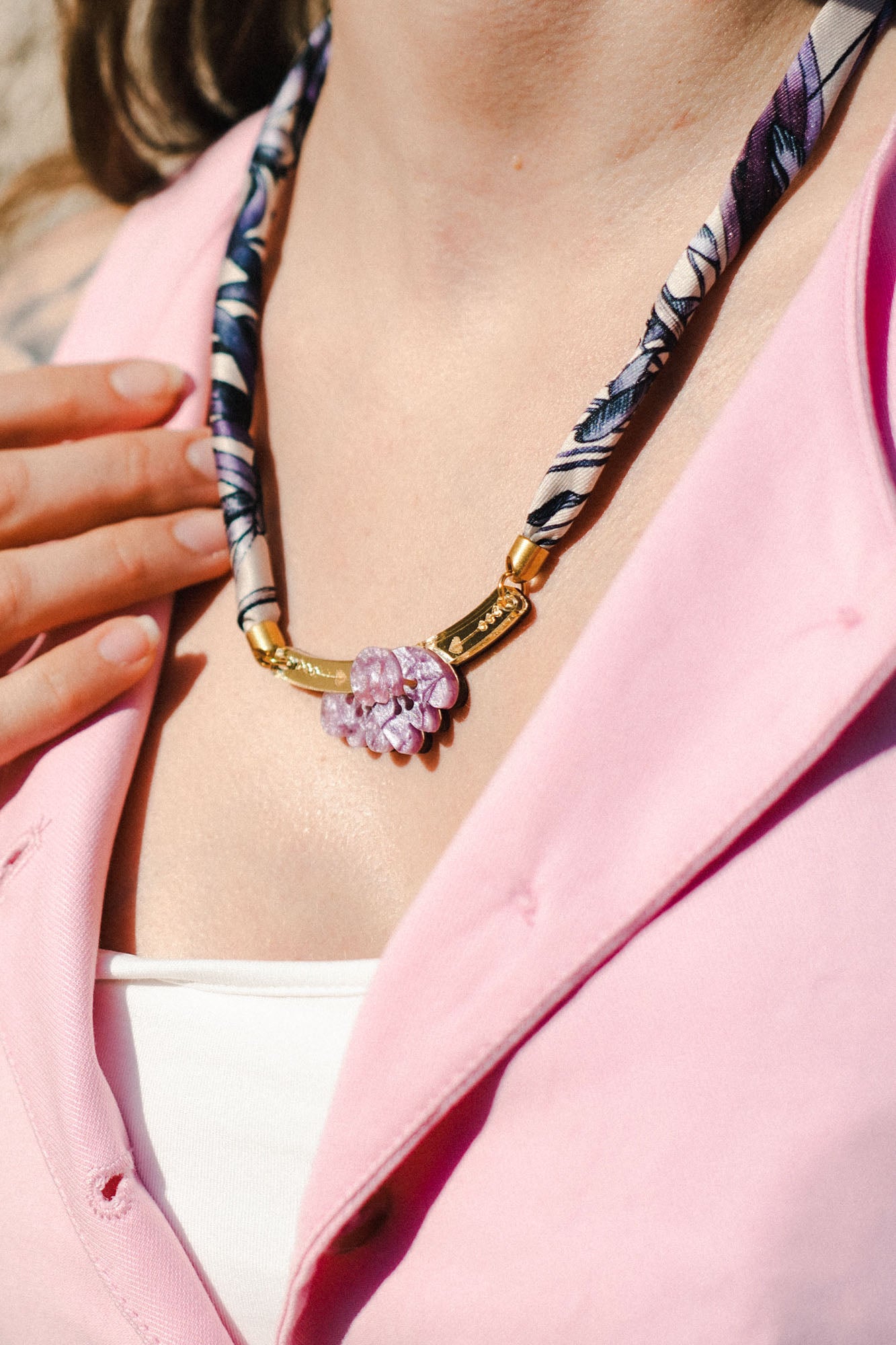 A person wearing a pink shirt and white top displays a handmade necklace with a patterned fabric band and gold clasp, featuring pink and purple gemstone flowers. Their hand rests near the collarbone, complementing an elegant silk-print scarf.