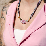 A woman wearing a light pink vest over a white top showcases a handmade necklace with a pink flower pendant; her arm features visible tattoos.