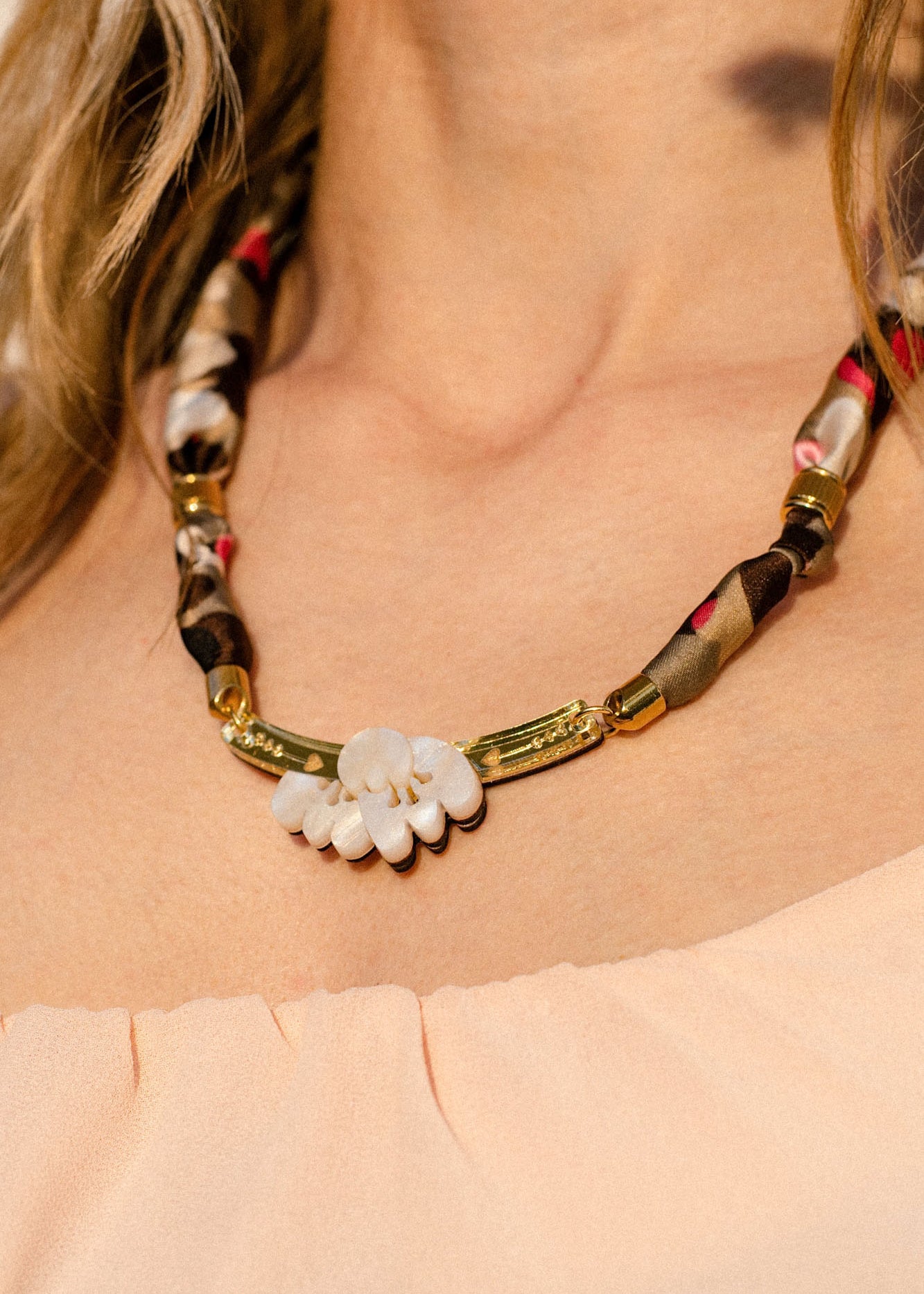 A close-up of a woman wearing a handmade necklace featuring an acrylic tulip pendant, gold accents, and an upcycled fabric-wrapped chain in brown, beige, and red tones; she is dressed in a peach off-shoulder top.