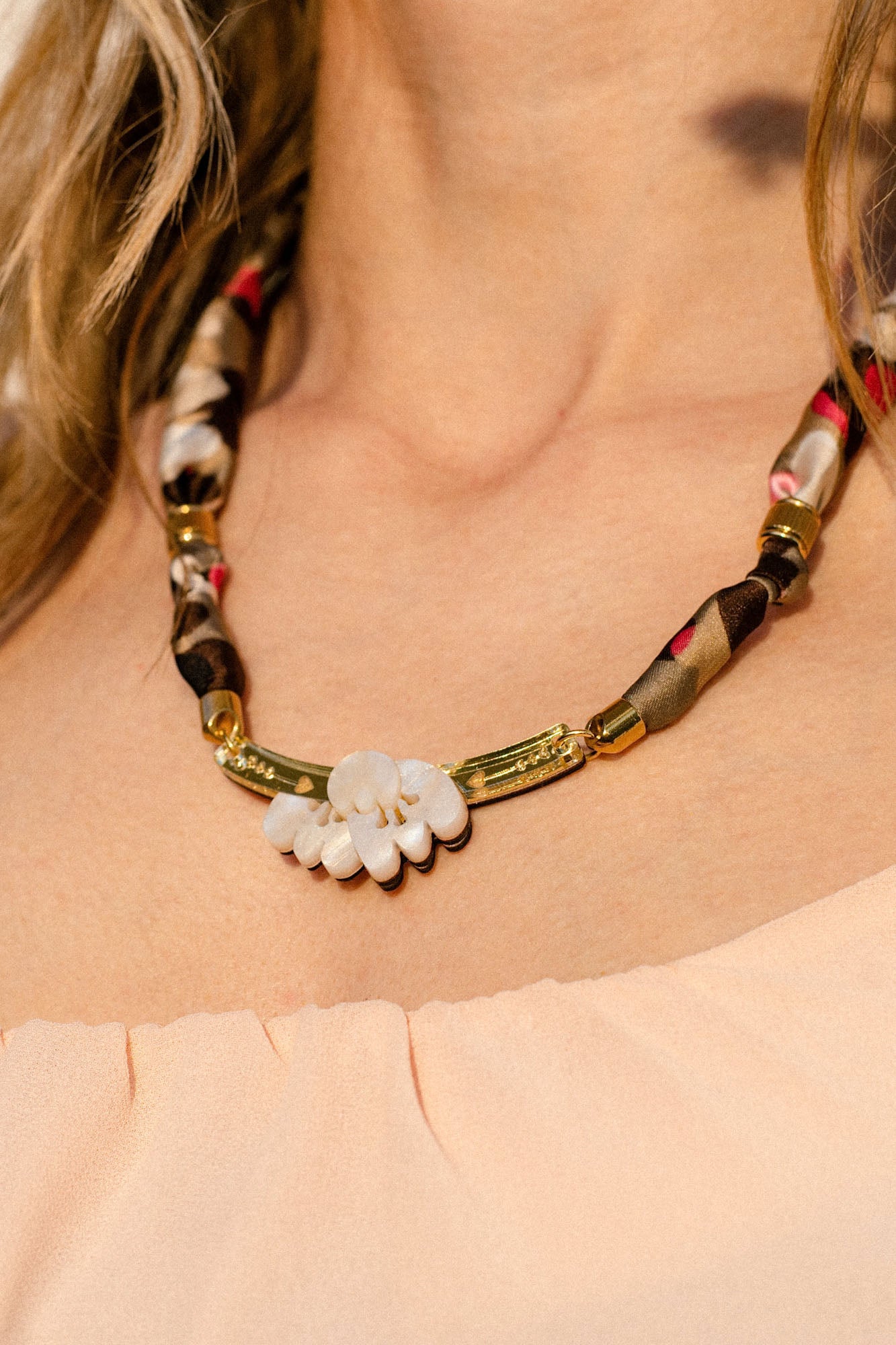 A close-up of a woman wearing a handmade necklace featuring an acrylic tulip pendant, gold accents, and an upcycled fabric-wrapped chain in brown, beige, and red tones; she is dressed in a peach off-shoulder top.
