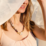 A woman wearing a pale pink sleeveless top and a handmade necklace smiles while partially hidden behind a flowing white fabric in soft sunlight.