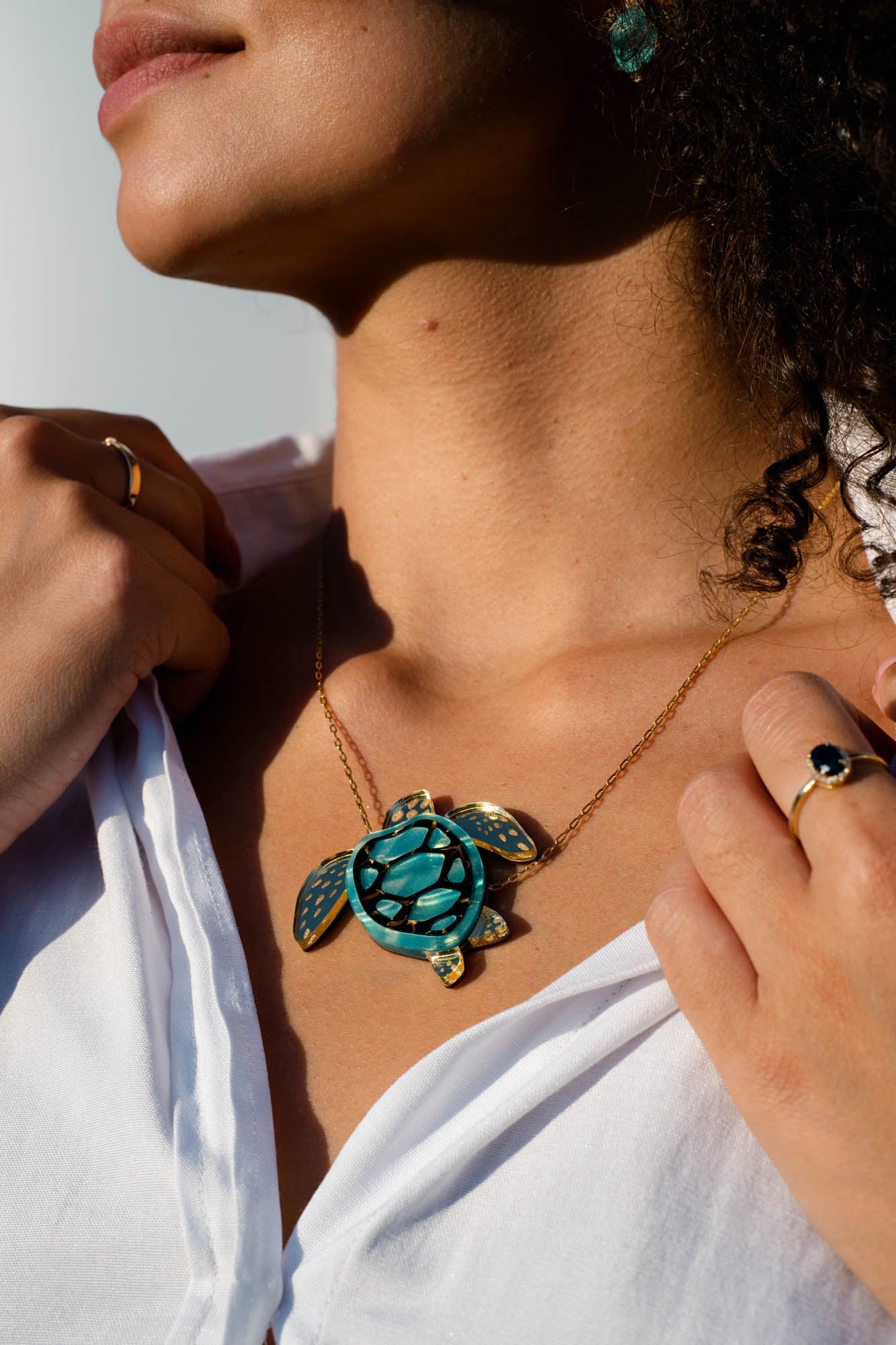 A woman wearing a white shirt displays a handcrafted Sea Turtle Necklace featuring a gold chain and a turquoise turtle pendant. She holds her shirt collar with both hands, showing rings on her fingers. Sunlight highlights her jewelry and skin.